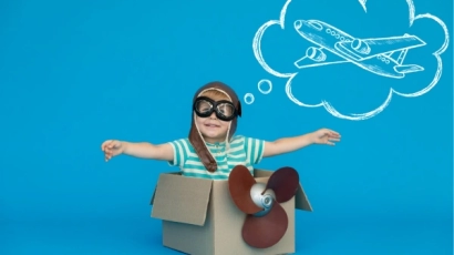 A child sitting in a cardboard box with a plastic propellor attached on its front. He is wearing aviator goggles and above him is a thought bubble with an aeroplane. Reading kids' stories broadens one's imagination.