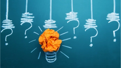 Picture of lightbulbs drawn over a chalkboard, with question marks in place of bulbs. The central lightbulb has a piece of crushed orange paper to illustrate light. This supports the concept of a strength of dyslexia being non-linear thinking.
