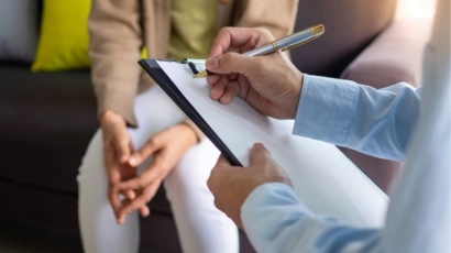 Psychologist writing down an observation on their clipboard, as the patient sits with their hands folded in the background. A professional must first observe the child to determine if they have special education needs