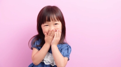 Girl smiling with her hands clasped over her mouth, against a blush pink background