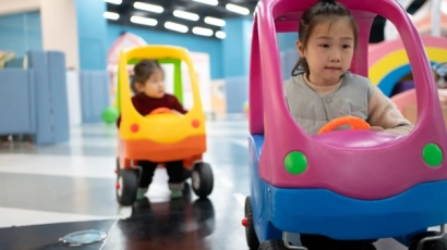 Children driving toy cars -- a girl in a pink and blue car in the foreground, and a boy in the yellow car in the background. VroomTown, one of the kids' indoor playgrounds on this list, offers a similar opportunity.