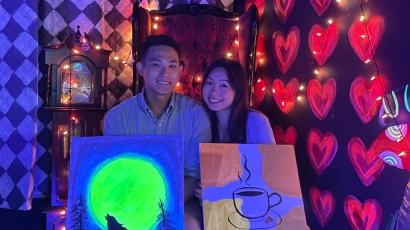 A couple posing in front of a wall of heart-shaped fairy lights, under blacklight. The man holds a portrait of a wolf howling at a neon green moon against a blue background, the woman holds a portrait of a coffee cup against a background consisting of abstract shapes in beige tones. They are posing at Scuro, which is one of the best kid friendly activities in Singapore.