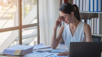 A woman pinching the bridge of her nose, looking exhausted, at a desk with papers spread all around her. Exhaustion is a symptom of caregiver burnout.