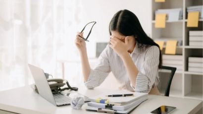 A woman with her head in her hands, in front of her desk with a stack of papers and a laptop. Feeling a lack of control over one's tasks is a contributing factor to caregiver burnout.