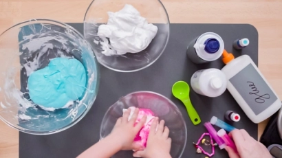Hands kneading slime in a glass bowl, with glue, saline solution and baking soda to their right. Slime is an excellent form of sensory play for toddlers.